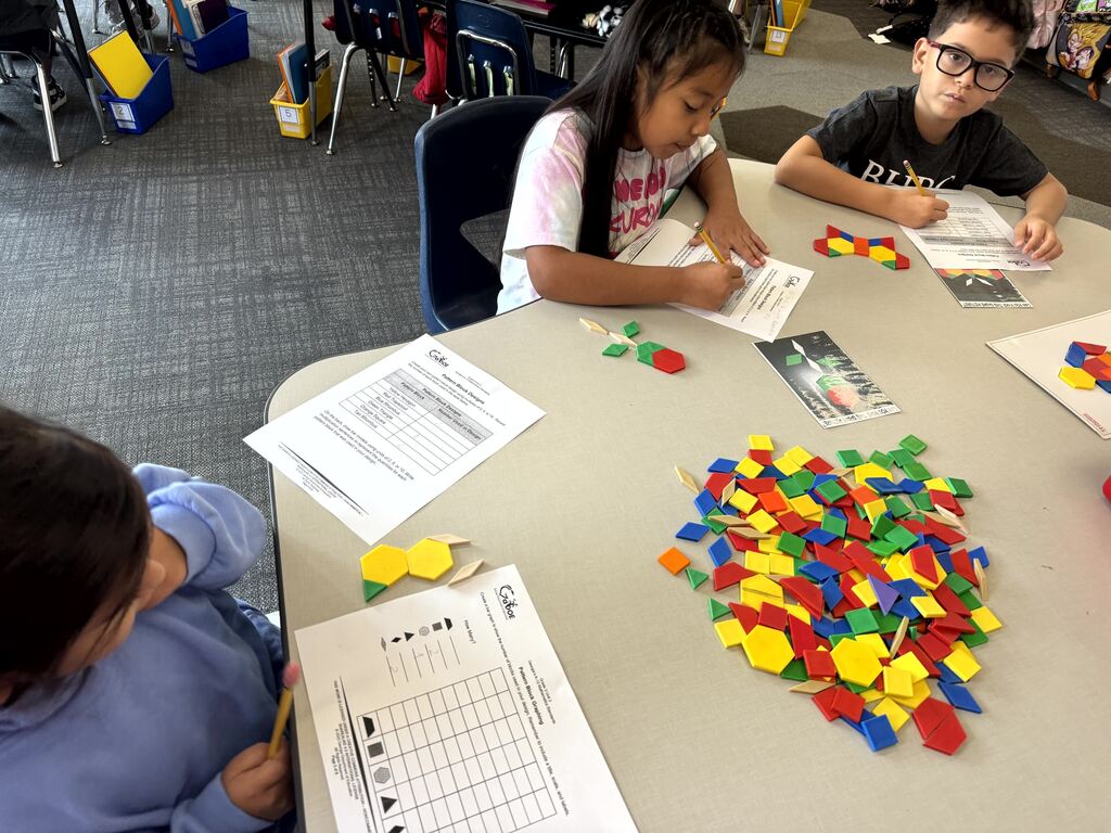 Short Alt Text: Three students sitting around a classroom table working with colorful plastic pattern blocks and math worksheets. Detailed Long Description: Three elementary students sit around a grey table working on a geometry assignment. A pile of multi-colored plastic pattern blocks (red hexagons, yellow trapezoids, blue rhombuses, green triangles) sits in the center of the table. Two girls and one boy write on worksheets labeled "Pattern Block Designs," arranging the blocks to create shapes and recording their work.