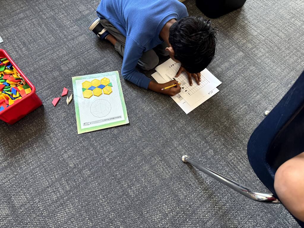 Activity: A student wearing a long-sleeved blue shirt kneels on a grey carpeted floor, leaning over a paper worksheet to write with a pencil. Materials On Floor: Worksheet: A grid graph where the student records the count of different geometric shapes. Template: A laminated sheet alongside the student featuring yellow hexagon and blue rhombus pattern blocks arranged in a symmetrical pattern. Storage Bin: A red plastic tub filled with colorful plastic pattern blocks (red, yellow, blue, green, and orange shapes) sitting to the left.