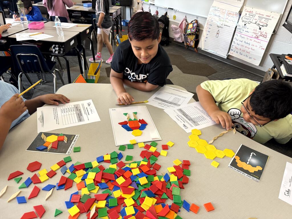 Activity: Two young boys are leaning over a light grey classroom table, focused on an educational math task using geometric shapes. Student Actions: The boy in the center (wearing a black t-shirt) smiles as he arranges colored pattern blocks onto a design template. The boy on the right (wearing a pale yellow shirt) leans close to the table, using his hands to align a string of yellow blocks. Materials On Table: A large, colorful pile of red, yellow, green, and blue plastic pattern blocks dominates the foreground. Surrounding the pile are activity instruction sheets, reference cards displaying various shape designs, and worksheets with blank graphing grids.