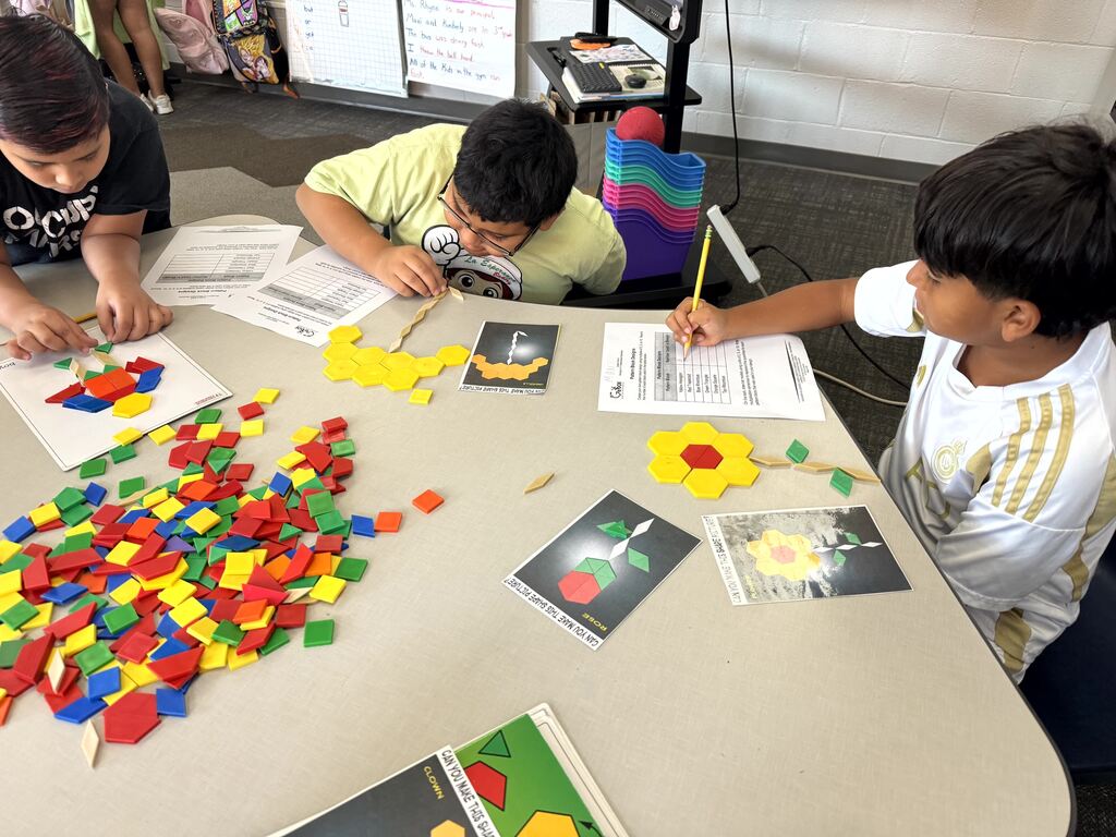 Three boys sit around a table filled with a large pile of colorful plastic pattern blocks and instructional activity cards. The students use yellow, red, green, and blue blocks to build flower and animal figures, writing down their answers on math worksheets with pencils.