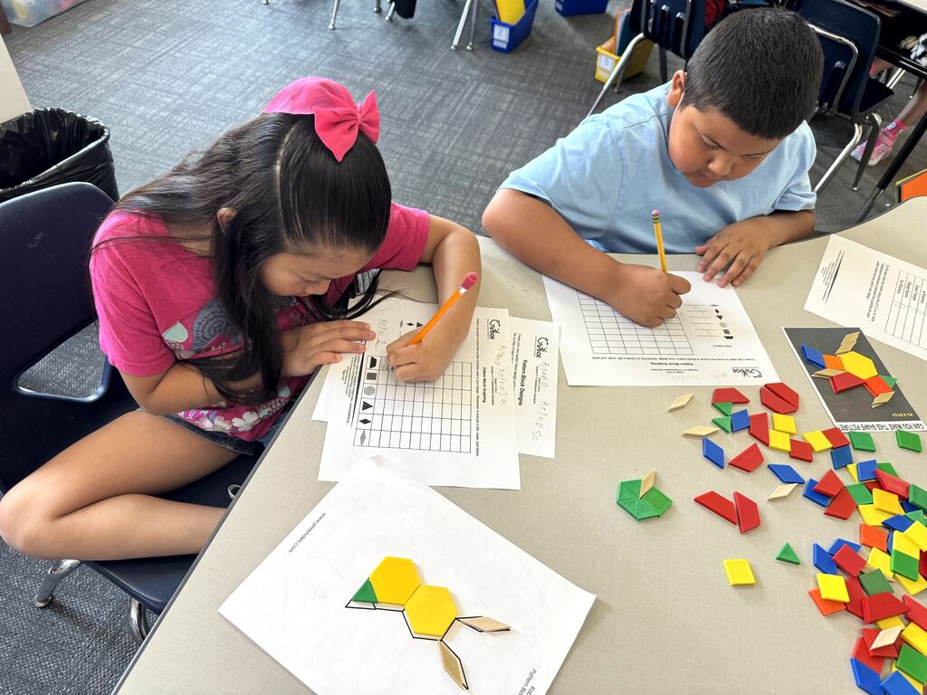 A girl with a bright pink bow in her hair and a boy in a blue shirt sit side-by-side at a classroom table, leaning over their worksheets and writing with pencils. A worksheet template displaying a yellow bunny made of pattern blocks lies in the foreground, with scattered plastic blocks nearby.