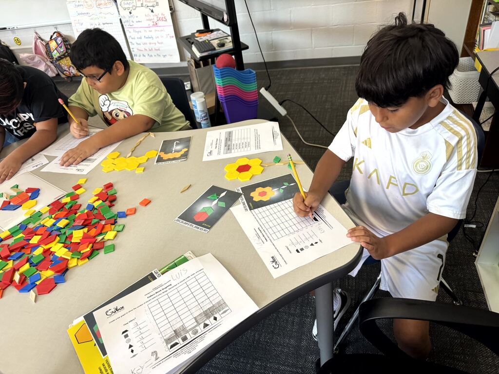 A student wearing a white Real Madrid soccer jersey sits at a curved table, using a pencil to write on a pattern block worksheet. On the table surrounding him are several geometric creations, including a yellow flower shape and various activity cards, next to a large pile of multi-colored pattern blocks.