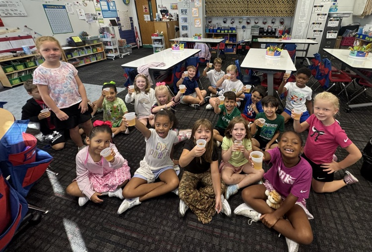 A group of elementary students sitting and kneeling on the classroom floor, proudly holding up small plastic cups filled with apple cider or juice toward the camera.