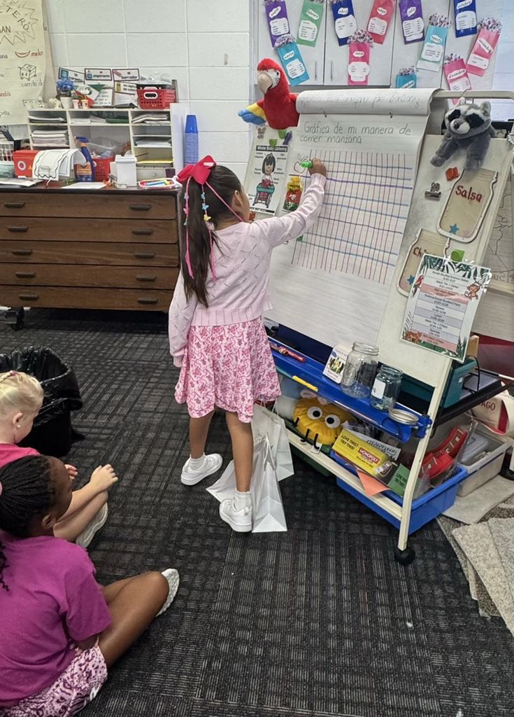 A young girl with long dark hair and a bright pink bow standing at a white easel, using a green marker to shade in a square on a chart titled "Gráfica de mi manera de comer manzana."