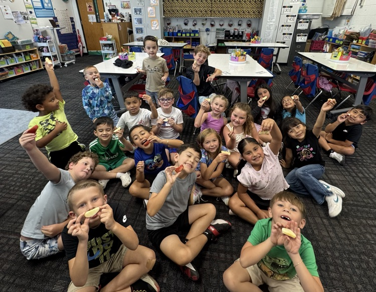 An overhead, wide shot of a group of young elementary students sitting cross-legged on a dark carpeted classroom floor, holding up fresh apple slices toward the camera.