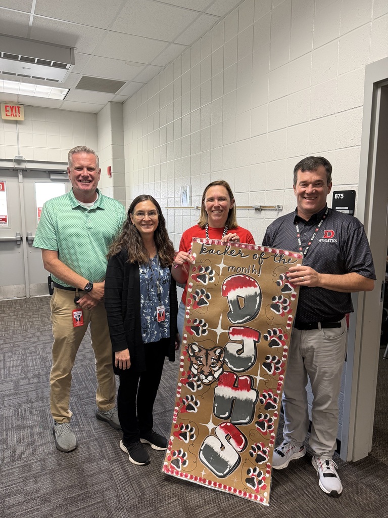 Four adults pose in a school hallway while holding a large handmade “Teacher of the Month!” sign featuring red, white, and gray school mascot illustrations, including paw prints, and a cougar..