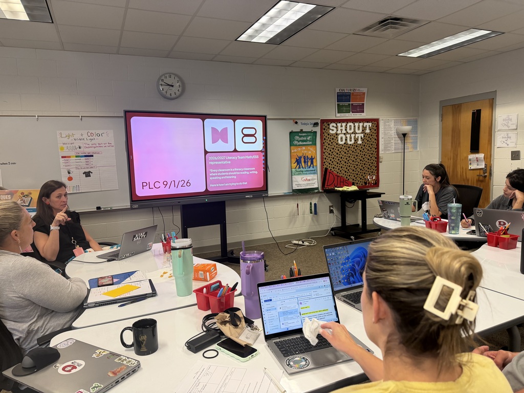 An over-the-shoulder view of a brightly lit school classroom or library space where several educators sit around white tables arranged in a circle for a professional development meeting. Open laptops, coffee mugs, water bottles, and school supplies rest on the tables. In the front of the room, a large digital monitor displays a slide titled "PLC 9/1/26" alongside the M8 logo and text about literacy team goals. Classroom whiteboards and posters, including a "SHOUT OUT" bulletin board, adorn the surrounding walls.