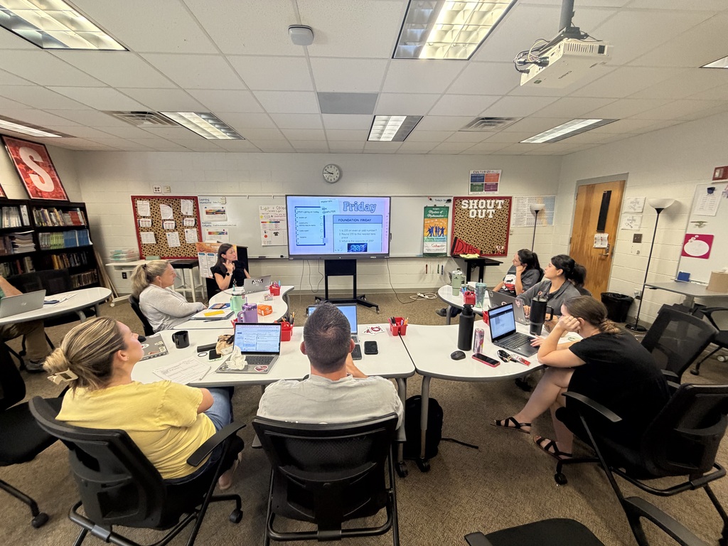 A wide-angle view shows a group of educators seated around white tables during a professional development meeting in a classroom. In the foreground, attendees sit facing a large digital display at the front displaying a slide titled "Friday." Open laptops, water bottles, and notebooks rest on the tables. Decorative whiteboards, a "SHOUT OUT" bulletin board, and bookshelves line the surrounding walls.