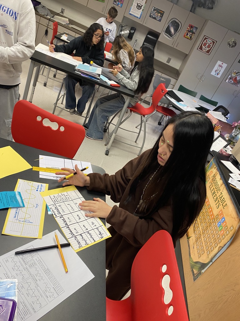 Student in the foreground sitting at a table working on science classwork. Students in the background doing the same.