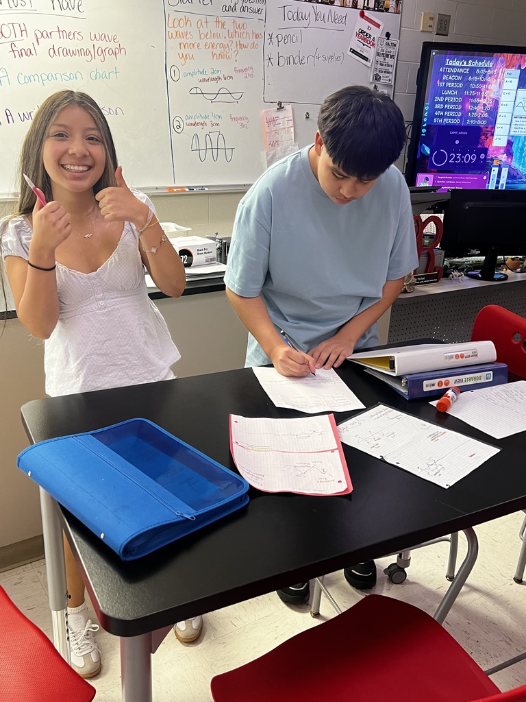 Two students in a science classroom. A female student in a whit shirt giving the "thumbs up" to the camera. The male student is writing on a paper on a black table.
