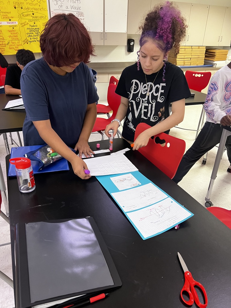 Two students in a science classroom working on a project at a black table with papers, glue sticks, and scissors.