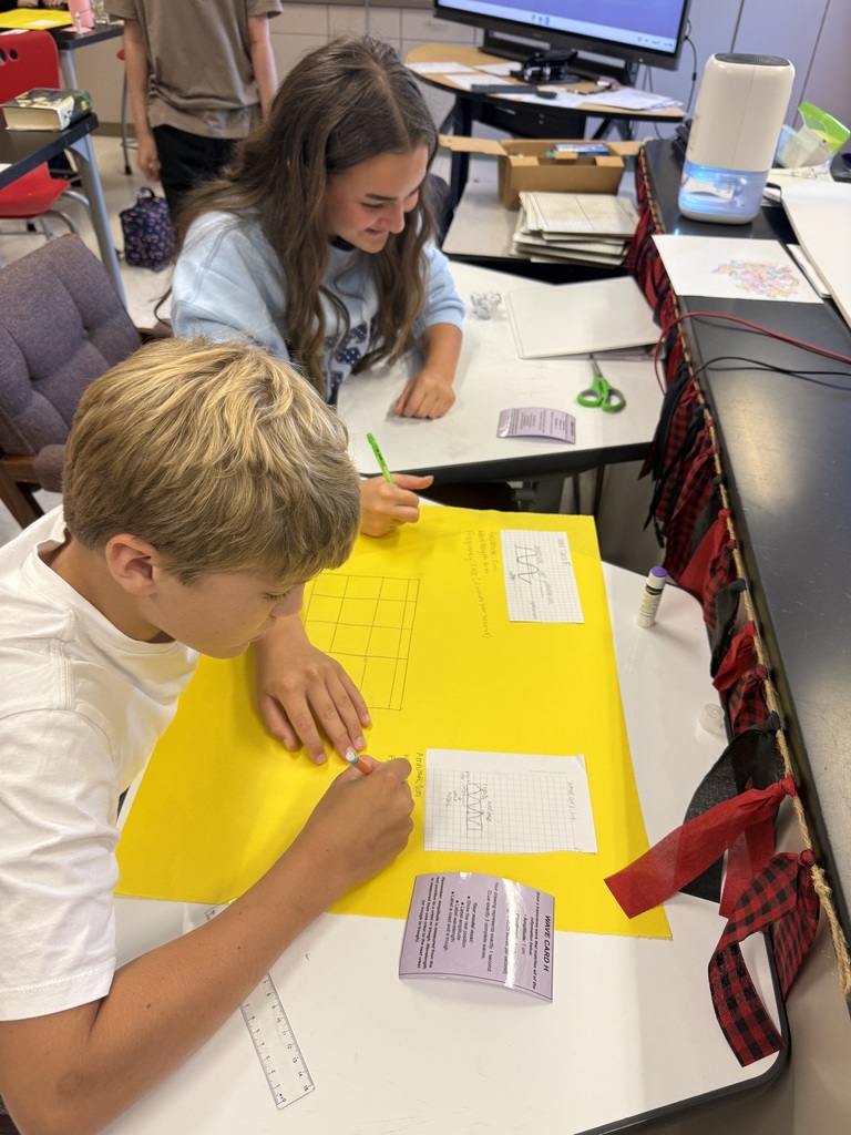 Two students in a science classroom working on a project on a yellow posterboard.