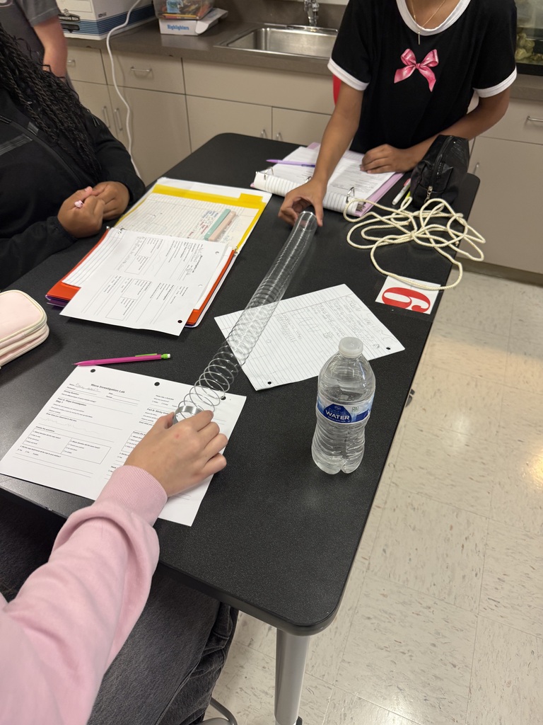 Students in a science classroom using a slinky to make waves. 3 students are sitting around a black table with papers, a water bottle, slinky, and rope on the table.