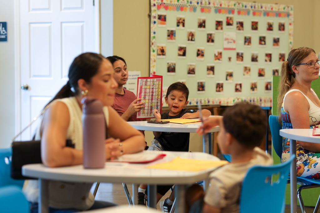 Multiple parents and young children sit at classroom tables reviewing paperwork. In the center, a mother holds an educational poster for her child, while other adults and children in the foreground look on attentively.