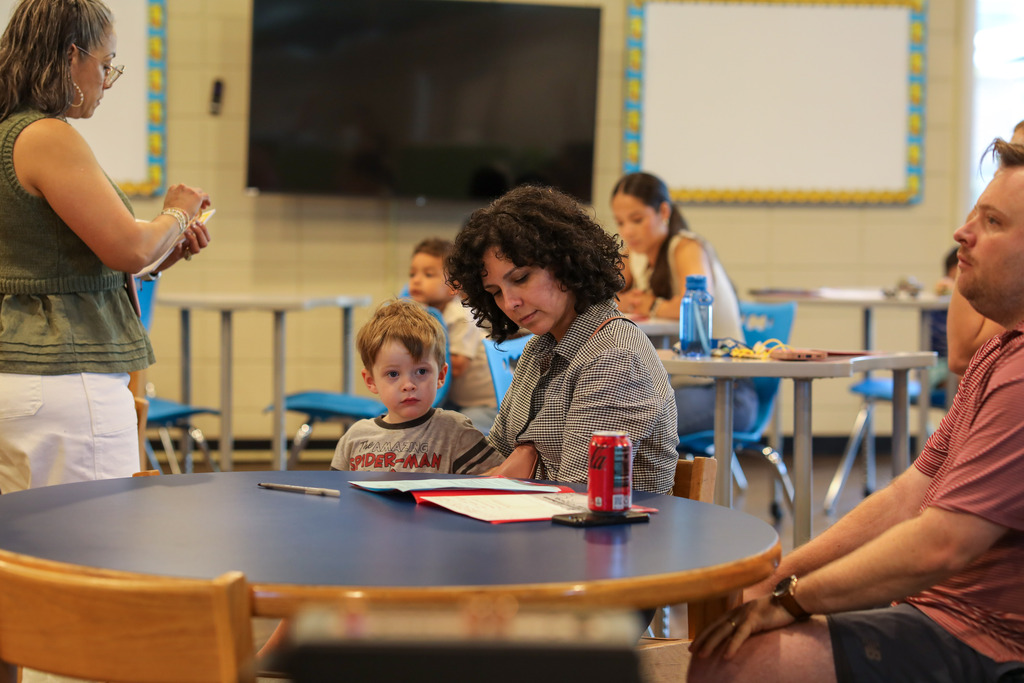 A woman with curly dark hair sits with a young child at a round blue table, reviewing printed forms alongside a red Coca-Cola can. A staff member in a green top stands nearby filling out papers.