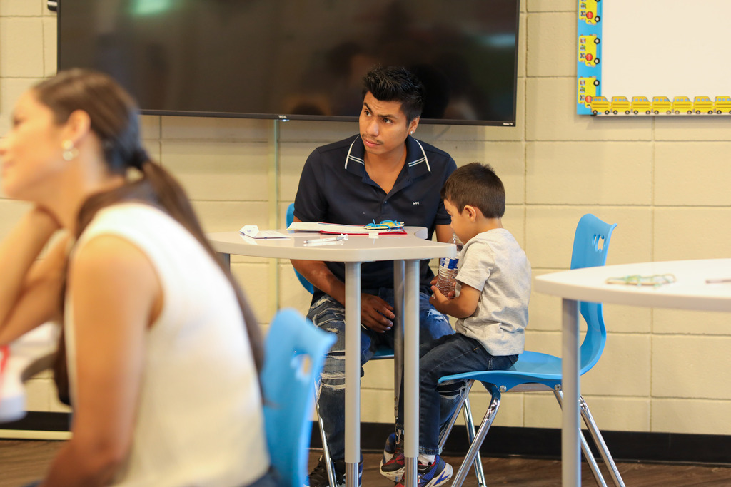 A man in a dark polo shirt sits at a gray table with a young boy wearing a gray shirt and jeans who holds a water bottle. In the foreground, a woman wearing a white top is seen partially out of focus, while classroom bulletin boards and a mounted TV display frame the background.