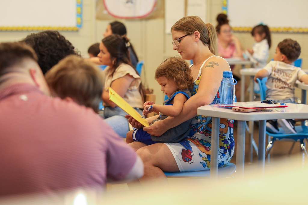 A woman with glasses holds a young child in a blue shirt on her lap as the child holds a pen and writes on a yellow folder. Other parents and children sit at surrounding tables in the brightly lit classroom environment.