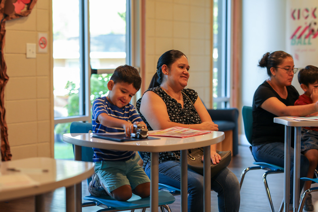 A young boy wearing a blue striped shirt plays with a small toy truck on top of a desk while his mother sits beside him smiling, with large windows providing natural light in the background.