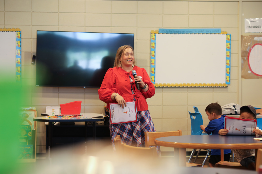 A woman in a bright red blouse and plaid skirt holds a microphone and a "Little Cats" booklet while addressing an audience in a classroom. In the foreground, young children sit at round tables holding matching orientation booklets.