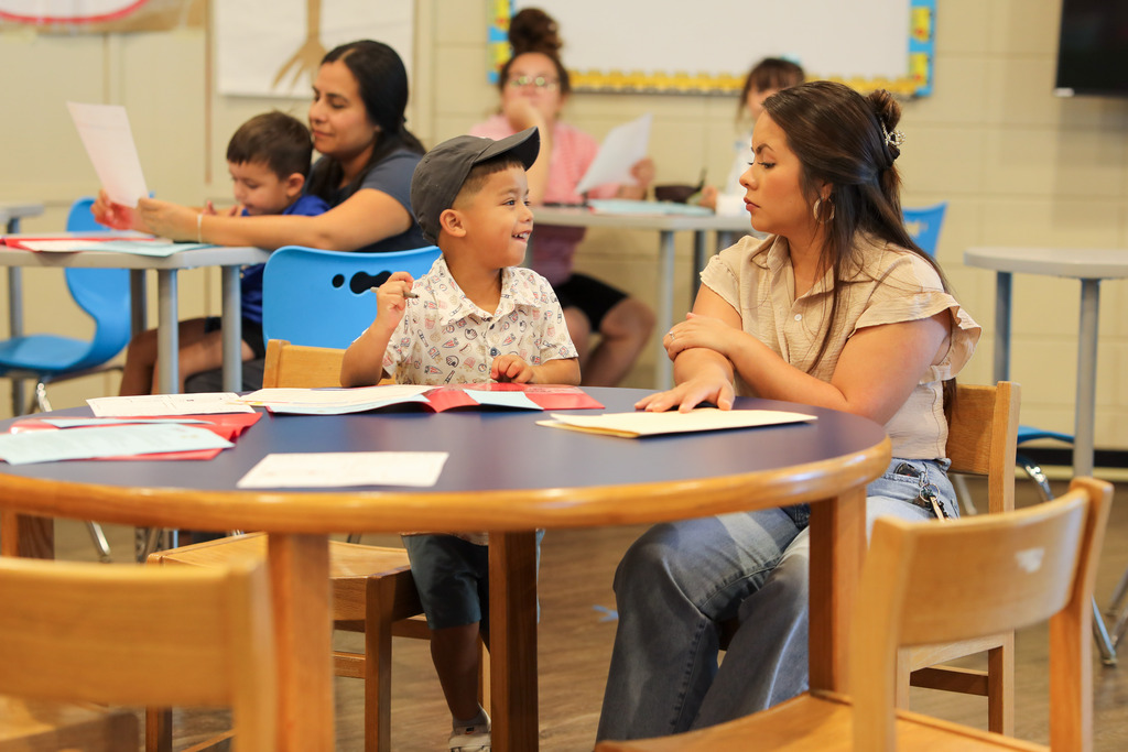 A woman in a beige blouse sits at a round wooden table, listening attentively as her young son, wearing a cap and patterned shirt, smiles and looks back at her while holding a pen over paper materials.