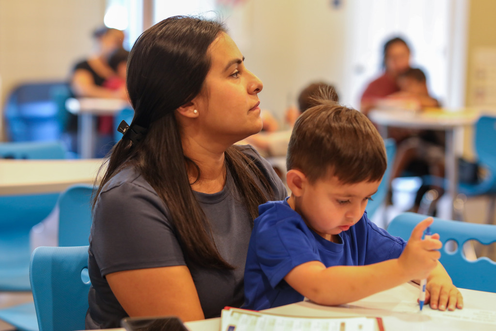 A mother looking ahead while her child writes on a paper on the desk.