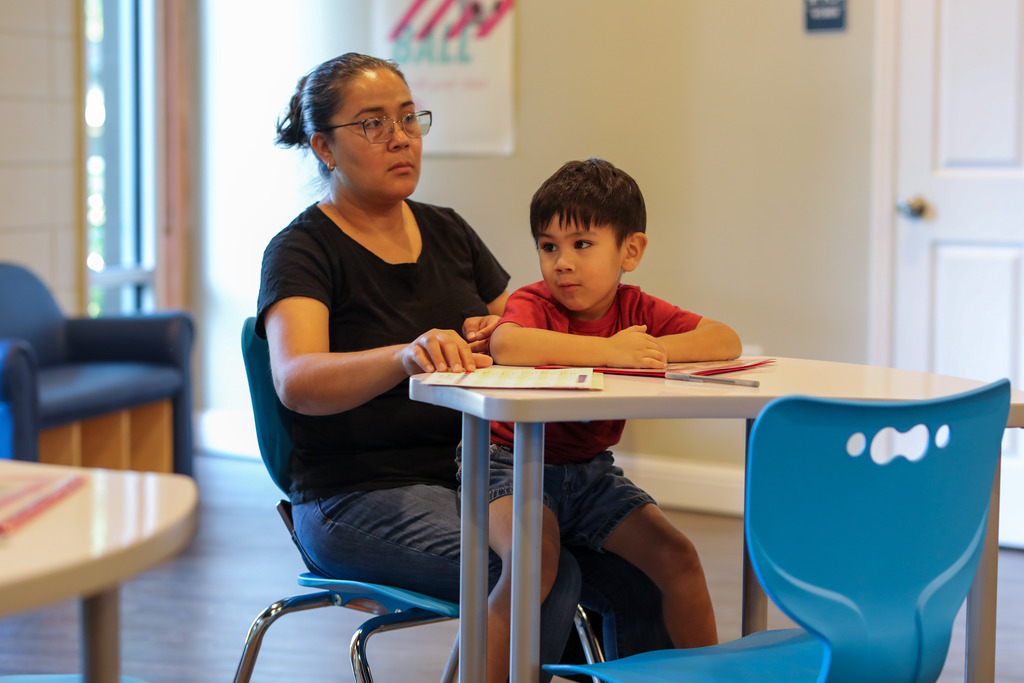 A woman wearing glasses and a black shirt sits beside her young son, who is dressed in a red t-shirt, at a small white classroom table with instructional materials laid out in front of them.