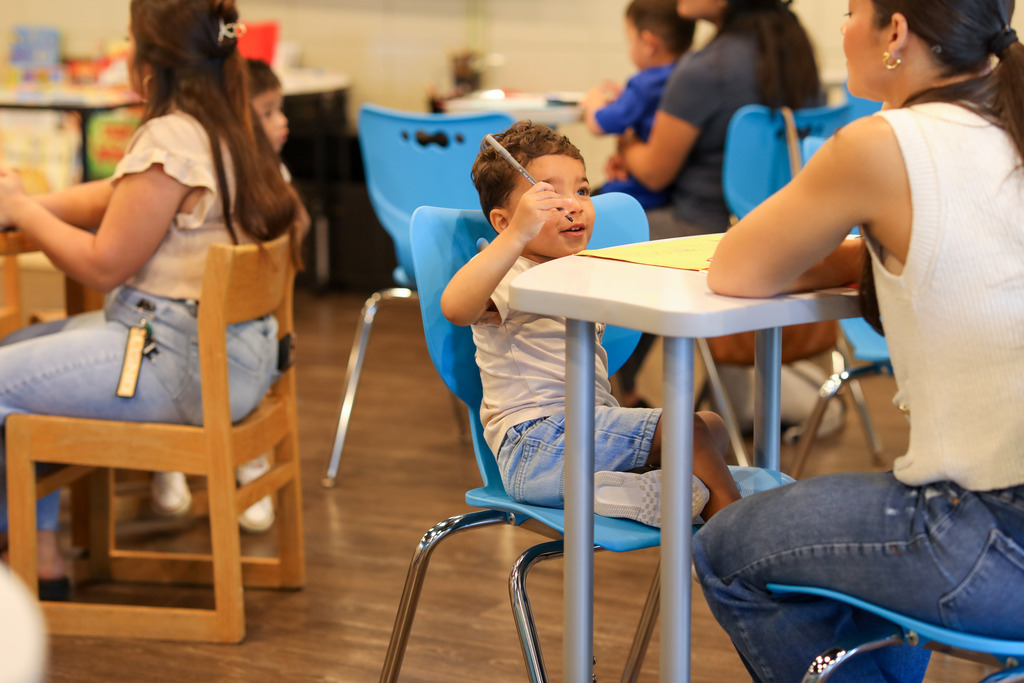 A toddler boy sitting in a light blue classroom chair holds a marker up toward the air, looking toward an adult sitting beside him at the desk. In the background, other families sit at desks participating in activities.