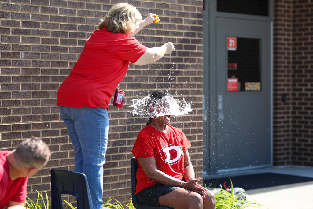 A woman standing on a chair pours water over a seated staff member's head, creating a dramatic freeze-frame splash of water covering the person's face.