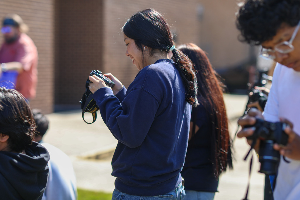 A female student wearing a navy sweatshirt stands outdoors smiling as she looks down at the preview screen of her Nikon DSLR camera. Other students around her hold cameras and focus on an event in the background.