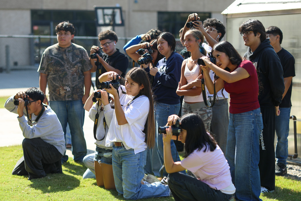A group of approximately ten high school students pose in various positions—crouching, kneeling, and standing—on a lawn next to a greenhouse as they align their camera lenses toward an outdoor activity.