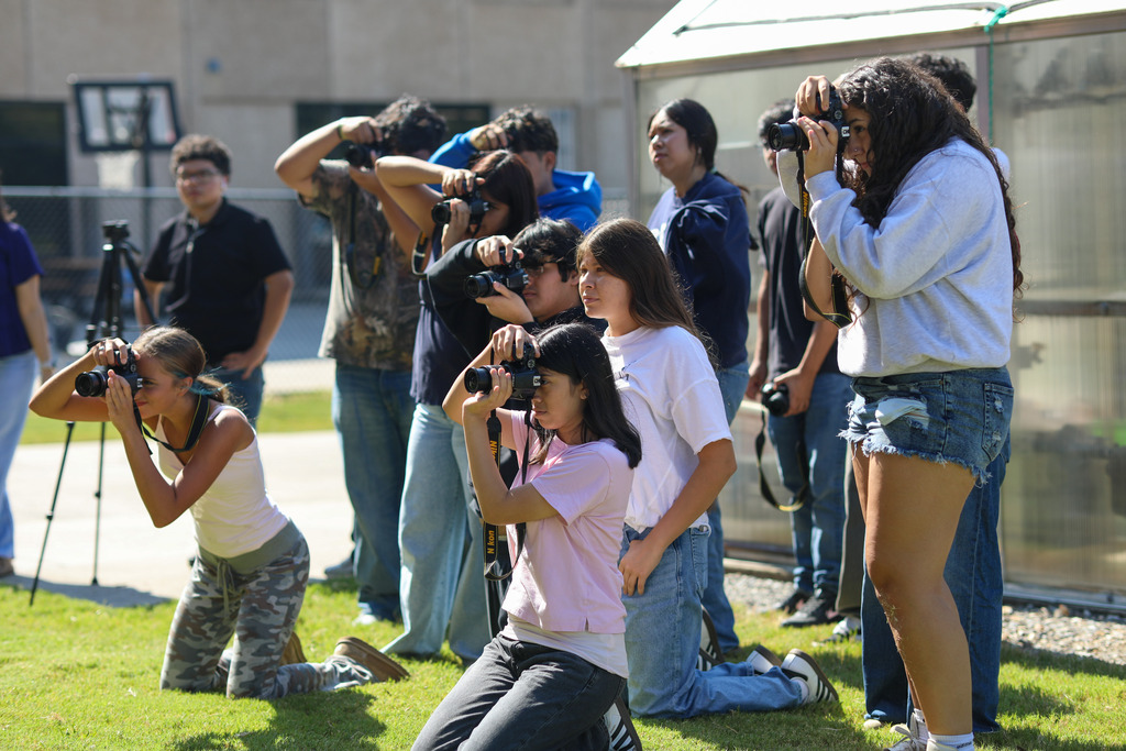 A group of student photographers stands and kneels on an outdoor lawn near a greenhouse structure, focusing their cameras forward. Several students hold DSLR cameras up to their eyes at varying heights, practicing capturing action photography during a school activity.