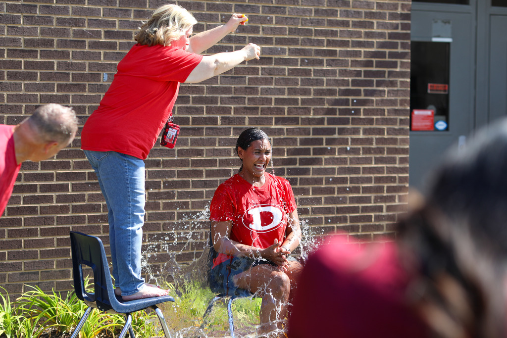 An outdoor school event scene where a woman in a red shirt stands on a plastic chair to squeeze or pour water directly over a seated colleague wearing a red Dalton shirt, capturing high-action water splashes.
