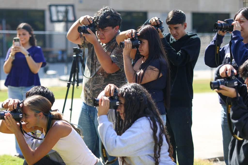 A group of high school students gather outdoors with DSLR cameras pressed to their eyes, aiming toward a subject off-camera. In the left background, a woman holds up her smartphone to record, while a tripod sits ready in the middle ground.