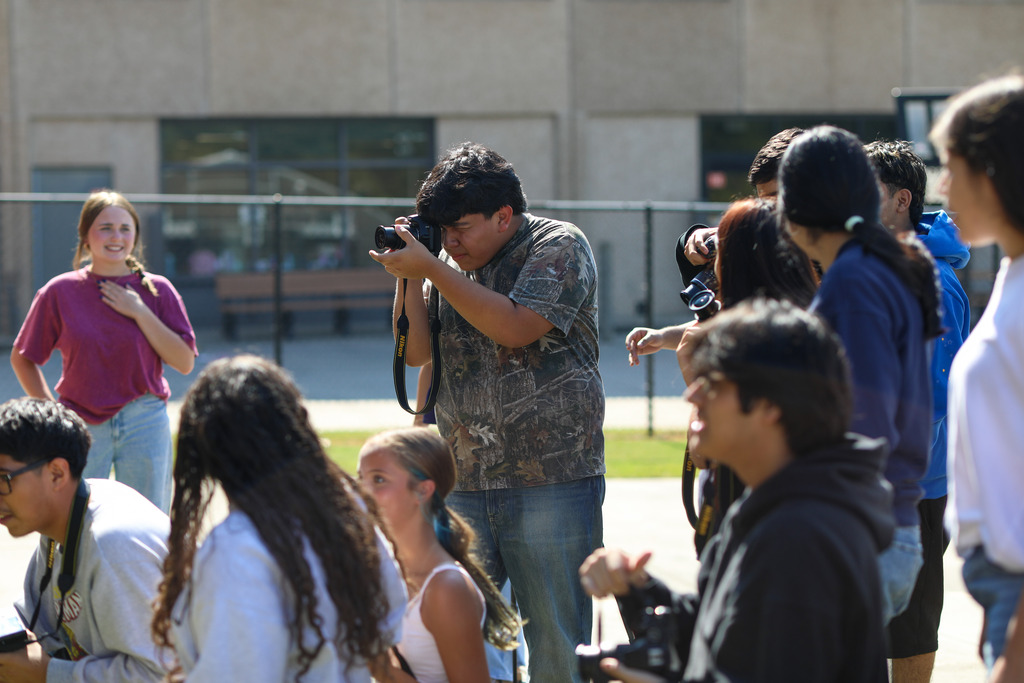 A male high school student wearing a camouflage shirt focuses his DSLR camera in a crowd of peers during an outdoor photography lesson or event, while other students around him prepare their own cameras.