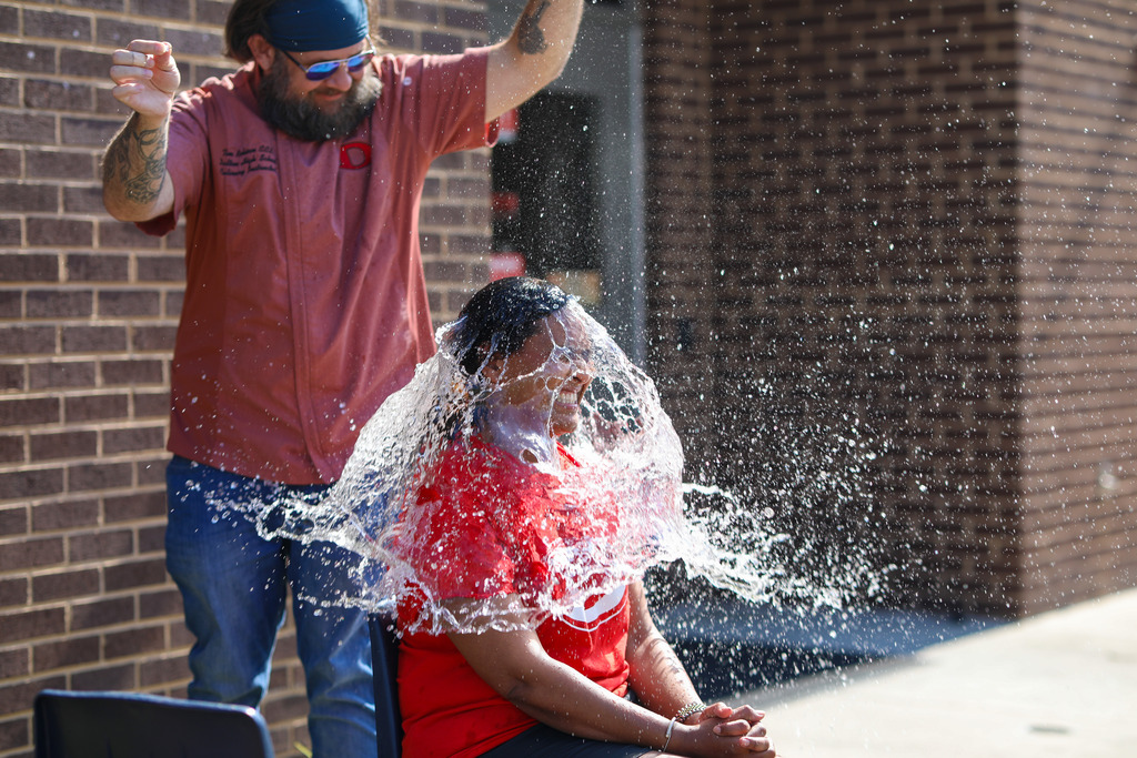 A male staff member with a beard and sunglasses reacts merrily as a large wave of water splashes over a seated colleague in a red shirt during an outdoor school activity.