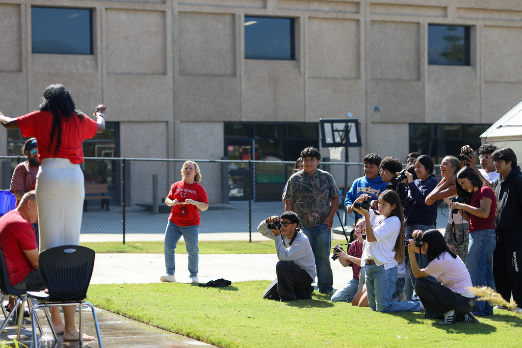 A wide-angle view showing a crowd of student photographers lined up on the grass holding cameras focused on staff members participating in a water activity in front of a brick school building.