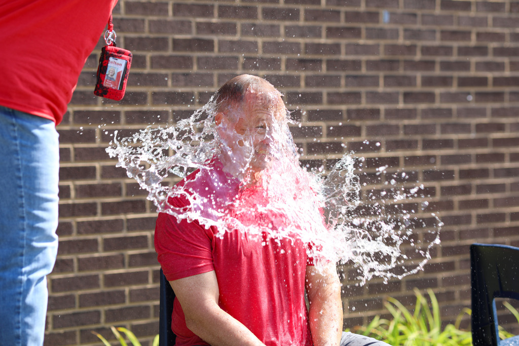 A close-up action photograph capturing the exact moment a bucket or sponge of water hits a seated male staff member's head and chest, creating a wide spray of water droplets against a brick wall background.