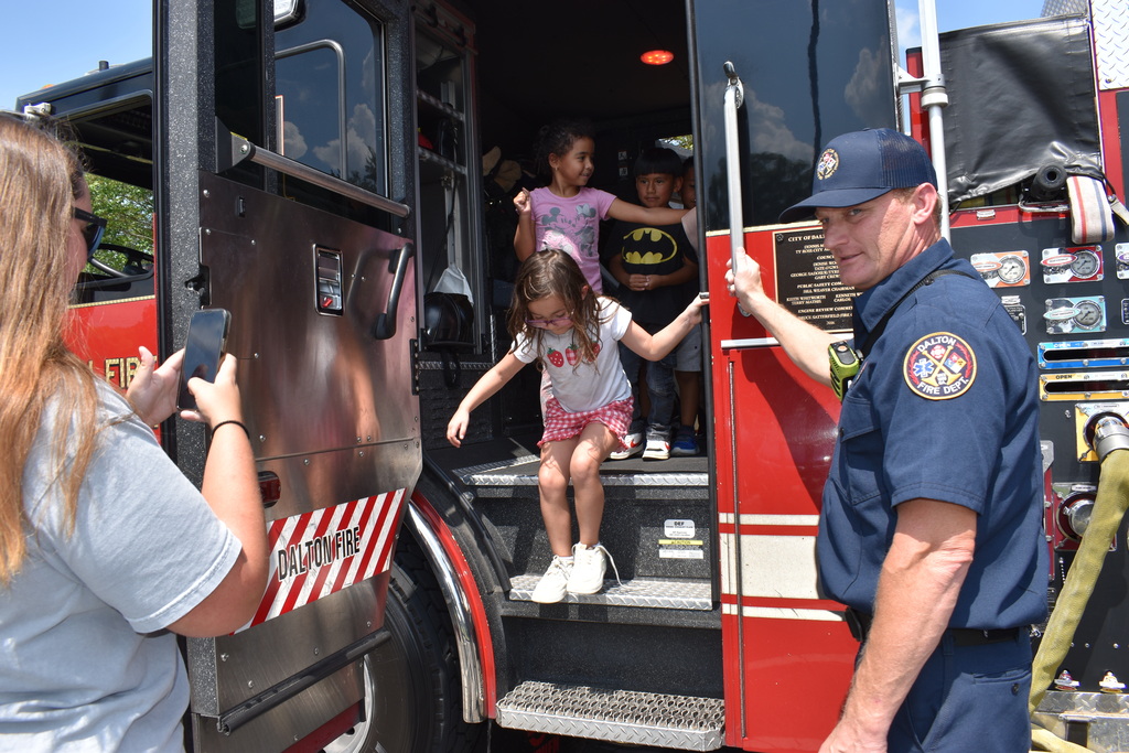 Main Subject: A young girl in a patterned shirt and pink shorts carefully steps down from the open door of a red fire truck.  Actions: A firefighter wearing a Dalton Fire Dept uniform and baseball cap stands beside the steps, monitoring her safety. Other children wait inside the cab doorway. In the foreground, an adult captures a photo with a smartphone.  Details: Visible markings on the fire truck read "DALTON FIRE".