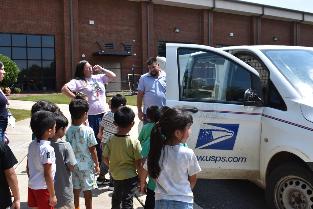 Main Subject: A group of young children stands on a sidewalk viewing a white USPS delivery van with its front door open.  People: A mail carrier in a light blue shirt stands near the vehicle door talking with the children, accompanied by adult supervisors standing nearby.  Details: The USPS emblem and website address ("usps.com") are prominently displayed on the side of the mail truck.