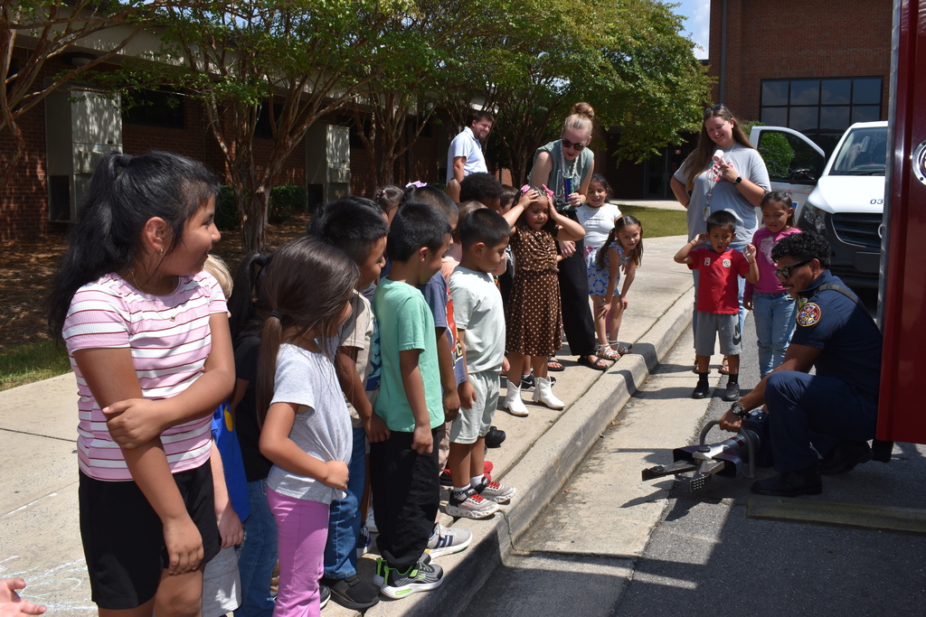 Main Subject: A firefighter crouches on the asphalt near a fire vehicle to demonstrate heavy hydraulic rescue equipment (spreader/cutter tool) to a group of children standing along the sidewalk curb.  People: A line of diverse elementary students watches attentively alongside two adult staff members supervising the group.