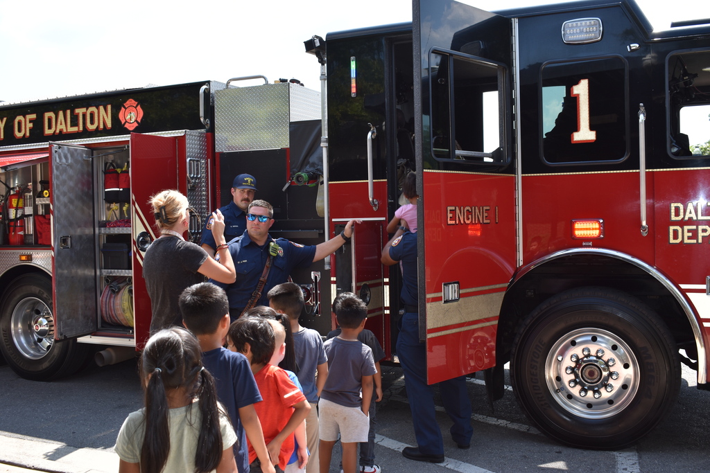 Main Subject: A group of young elementary students queue in a line alongside a red and black City of Dalton Fire Department Engine 1 firetruck.  Actions: A firefighter in uniform stands by the open door helping children enter the cab, while another firefighter interacts with the group. A teacher stands nearby taking photos with a smartphone.  Setting: Outdoors on a paved driveway/parking area under bright daylight.