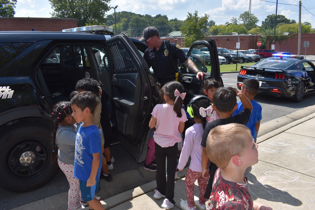 Main Subject: A police officer in uniform leans into the open door of a black police utility vehicle, explaining features to a group of young children clustered around.  Setting: Outdoors along a sidewalk. In the background, a sports car police cruiser with emergency lights active is parked in a parking lot.