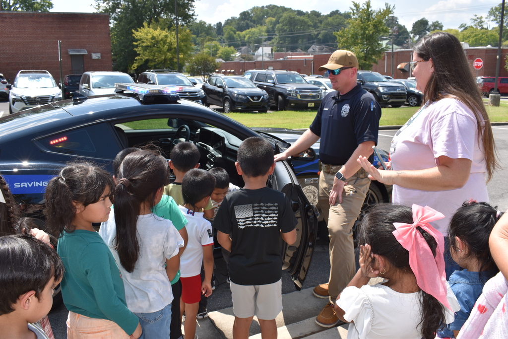 Main Subject: A police officer wearing a cap and sunglasses stands by the open driver-side door of a black police vehicle, talking to a gather of young students and a female teacher in a light pink shirt.  Details: The children are looking closely into the vehicle’s interior. Background shows a parking lot with several parked cars under a sunny sky.