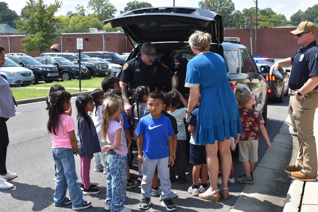 Main Subject: A police officer reaches inside the open rear hatch of a black police SUV while children gather closely to look inside.  People: A female teacher in a blue dress stands with the children, and a second officer in a cap and sunglasses stands nearby directing attention.  Setting: Outdoor parking area near a school building with parked vehicles in the background.