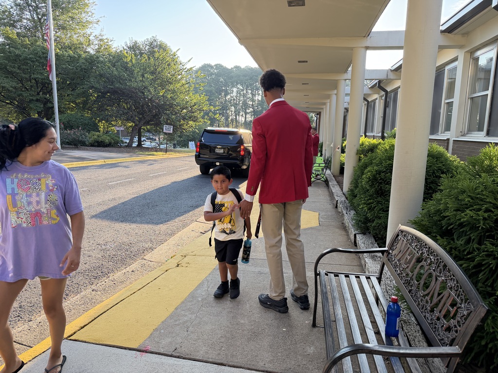 Student in a red jacket high-fiving a younger student as he walks inside the school