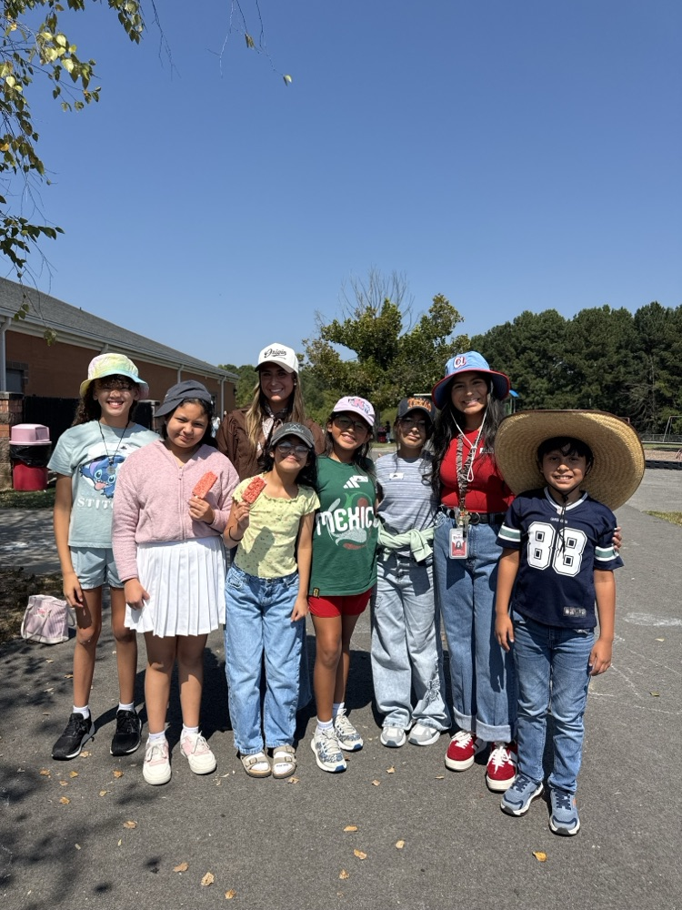 A group of eight students and adults standing outside in a paved school yard on a sunny day, posing for a group picture while wearing a variety of fun hats, including bucket hats, baseball caps, and a large wide-brimmed straw hat.