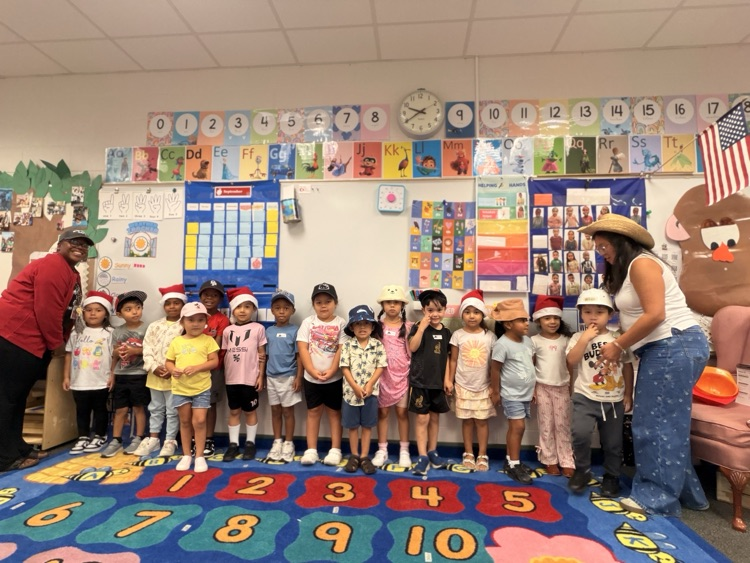 A row of young elementary school students and two teachers lined up on a colorful number carpet in a classroom, all wearing festive or fun hats such as Santa hats, bucket hats, and baseball caps.