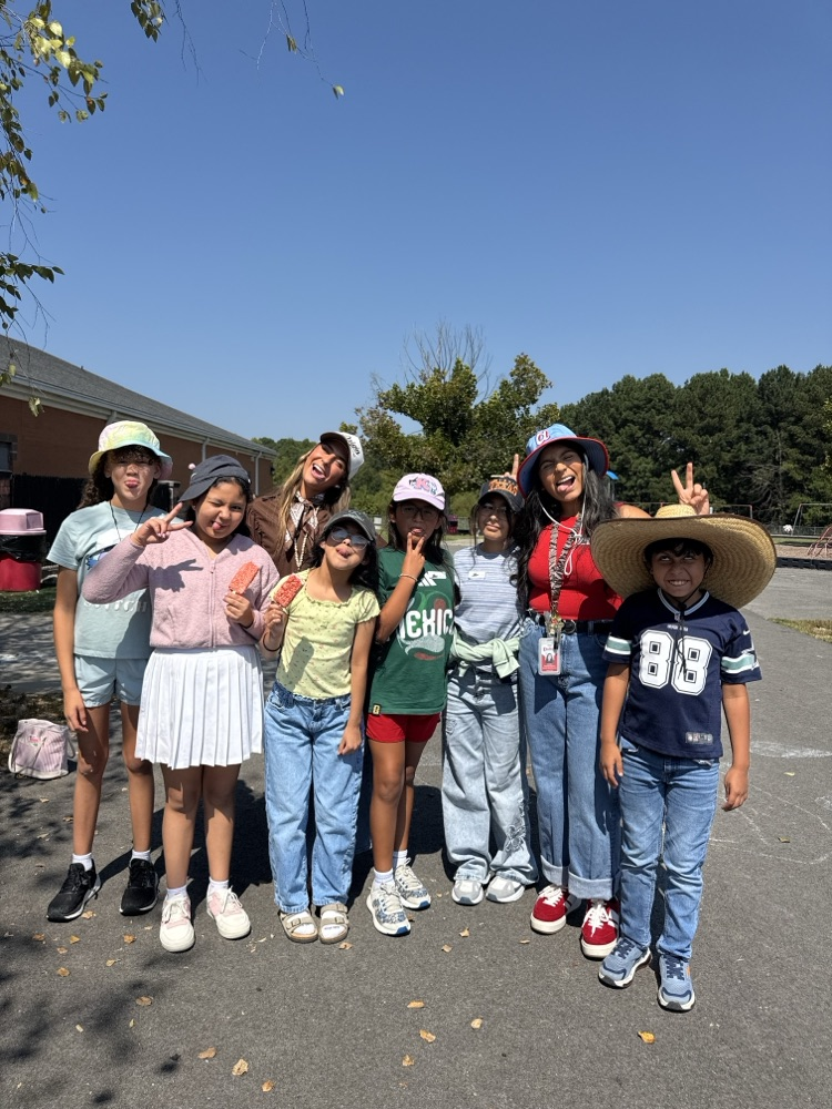 A cheerful group of students and adults posing together outside on a sunny day, making peace signs and fun faces while showing off their different headwear, including bucket hats, baseball caps, and a large straw hat.