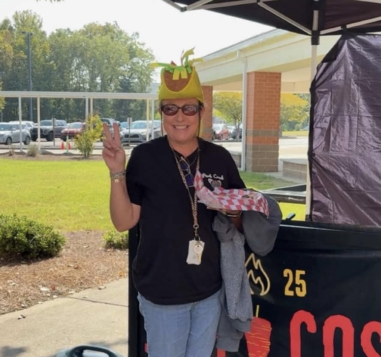 A smiling woman standing outdoors under an event tent, flashing a peace sign while wearing sunglasses and a novelty taco-shaped hat, holding a tray with food.