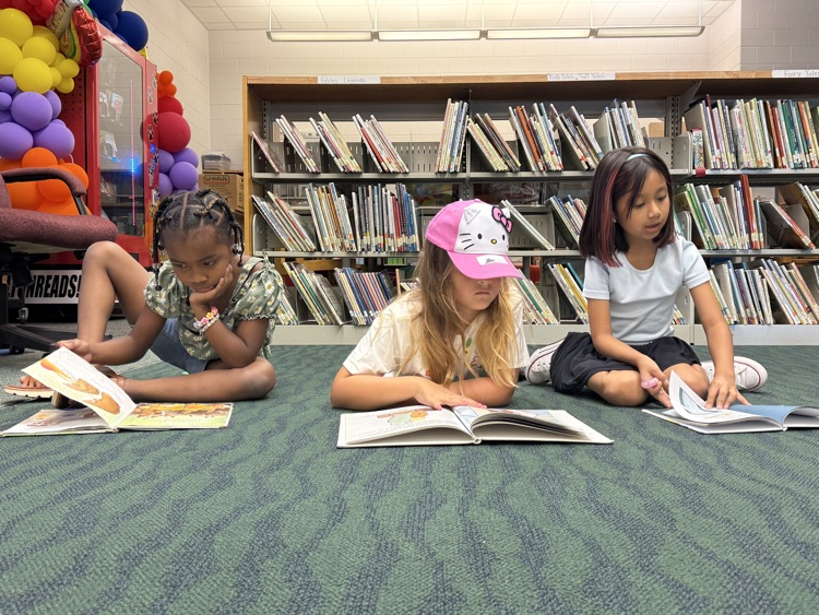 Three young students lying and sitting on the carpet floor of a school library, focused on reading open books, with one girl in the center wearing a bright pink Hello Kitty baseball cap.